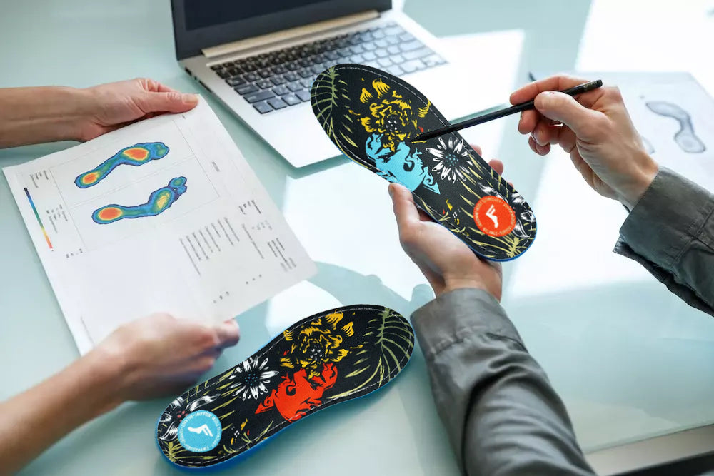 A photo of someone holding a custom orthotics insole in their left hand while pointing at the right one with a pencil, and a person to their left is holding a printout of a foot scan.