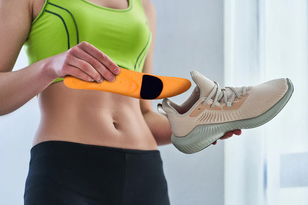 A fit woman wearing a green sports bra holds a running shoe in her left hand and is holding an arch support insole in her right hand ready to place it inside.